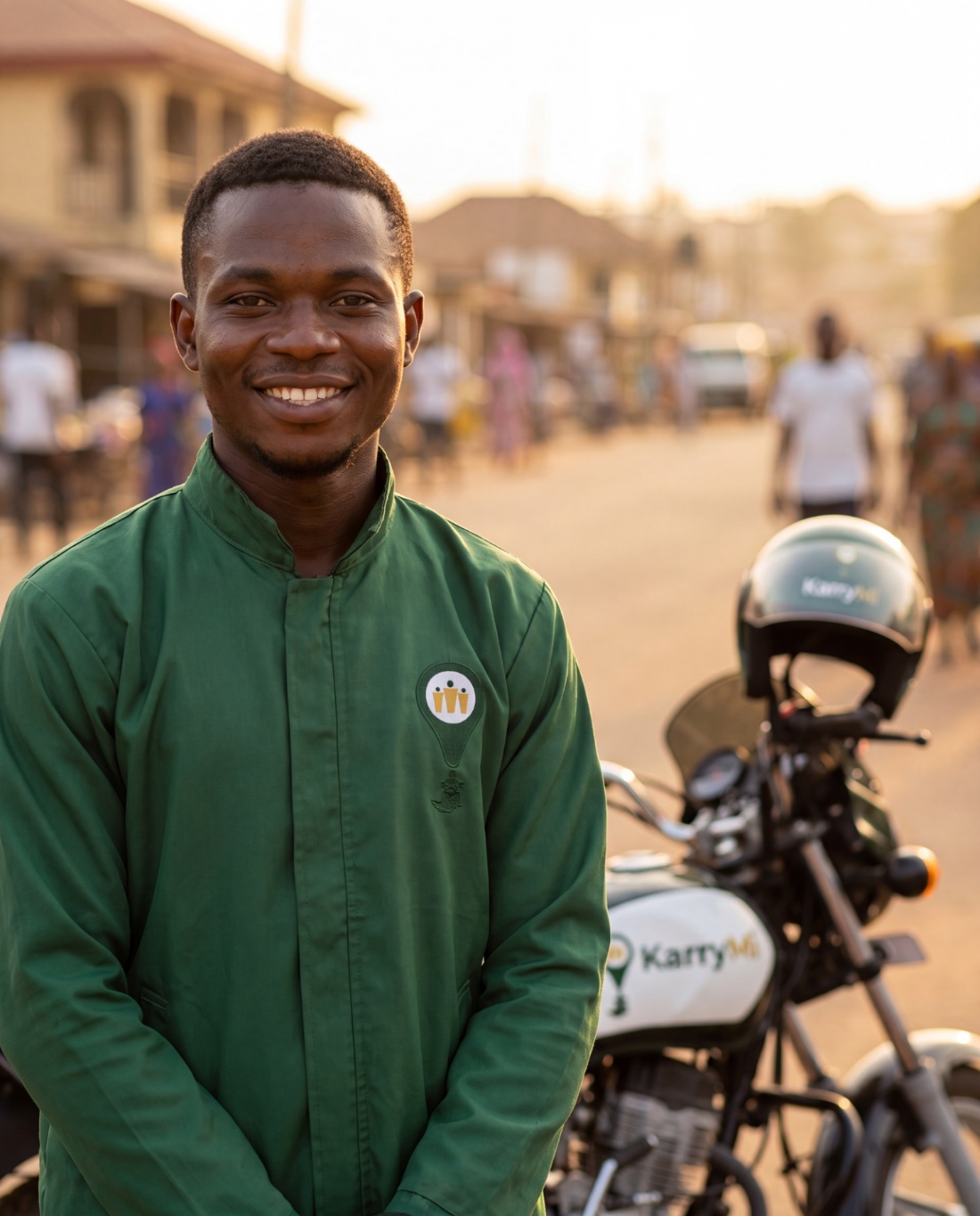 A KarryMi rider in a forest-green jacket smiling beside his branded motorcycle on an Ado-Ekiti street at golden hour